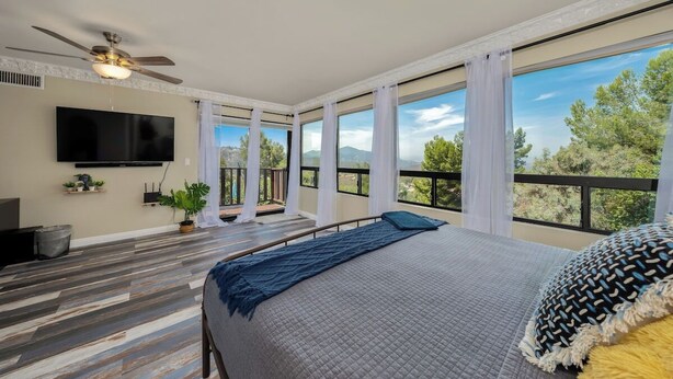 Une chambre dotée d’un grand lit, d’un ventilateur de plafond, d’un téléviseur fixé au mur et d’un balcon offrant une vue sur les arbres et les montagnes.