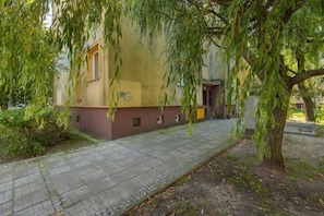 Un bâtiment avec un trottoir dallé, un arbre aux branches pendantes et un petit espace aménagé en jardin.