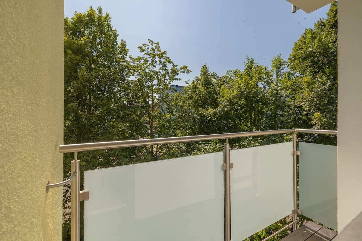Ein Balkon mit Metallgeländer und Glasbrüstung, der auf eine üppige, grüne Landschaft mit Bäumen blickt.