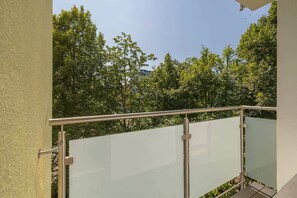 Ein Balkon mit Metallgeländer und Glasbrüstung, der auf eine üppige, grüne Landschaft mit Bäumen blickt.