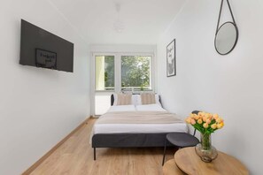 Une chambre moderne avec un lit, une table de chevet, un téléviseur fixé au mur et une fenêtre donnant sur des arbres.