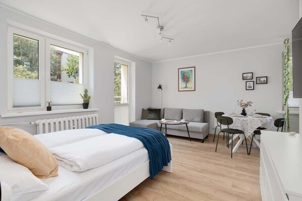 A modern bedroom with a bed, a gray sofa, a dining table with chairs, and a window with blinds.