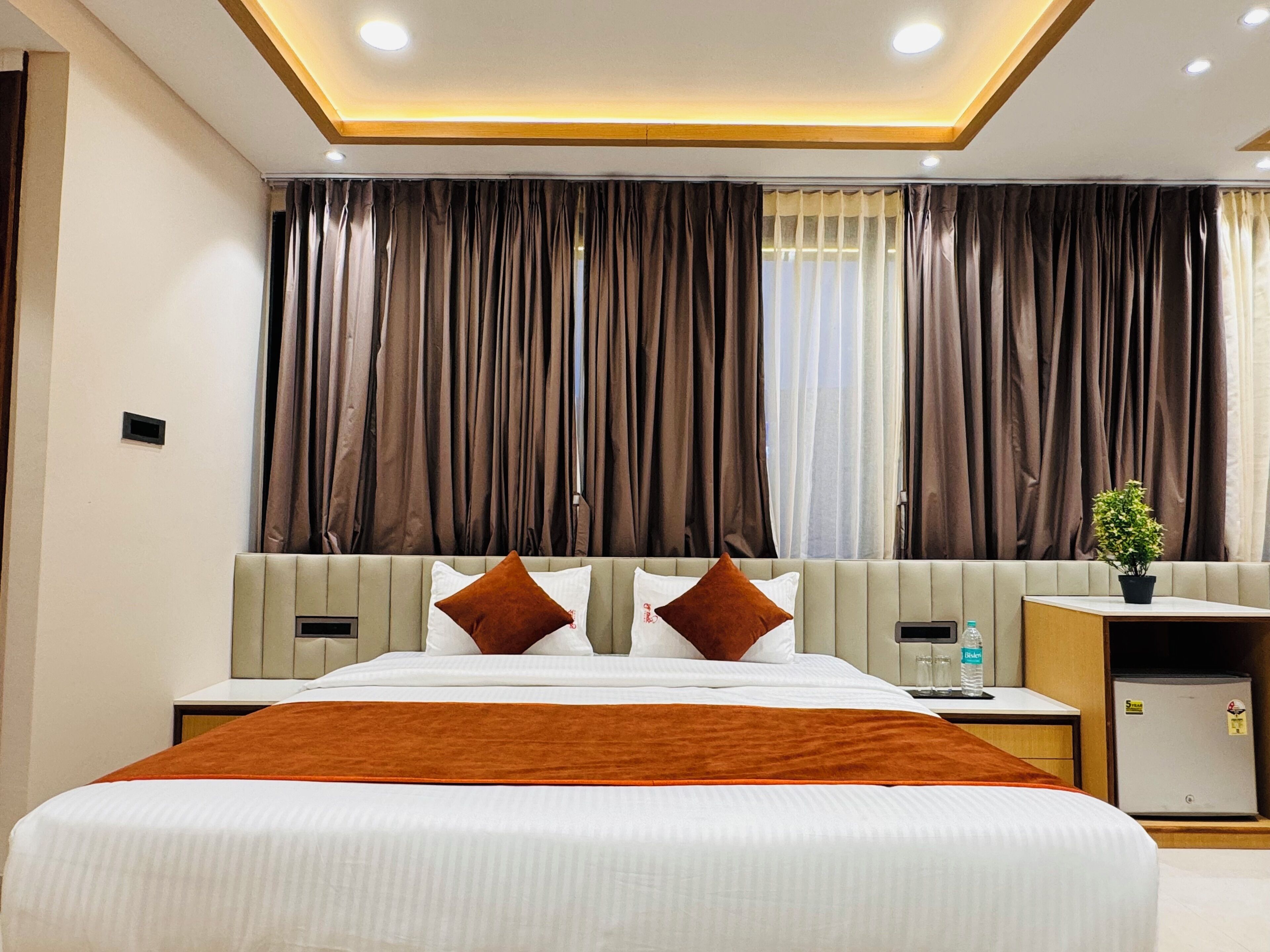 A hotel room with a large bed, a nightstand, a small table with a plant, and a refrigerator.