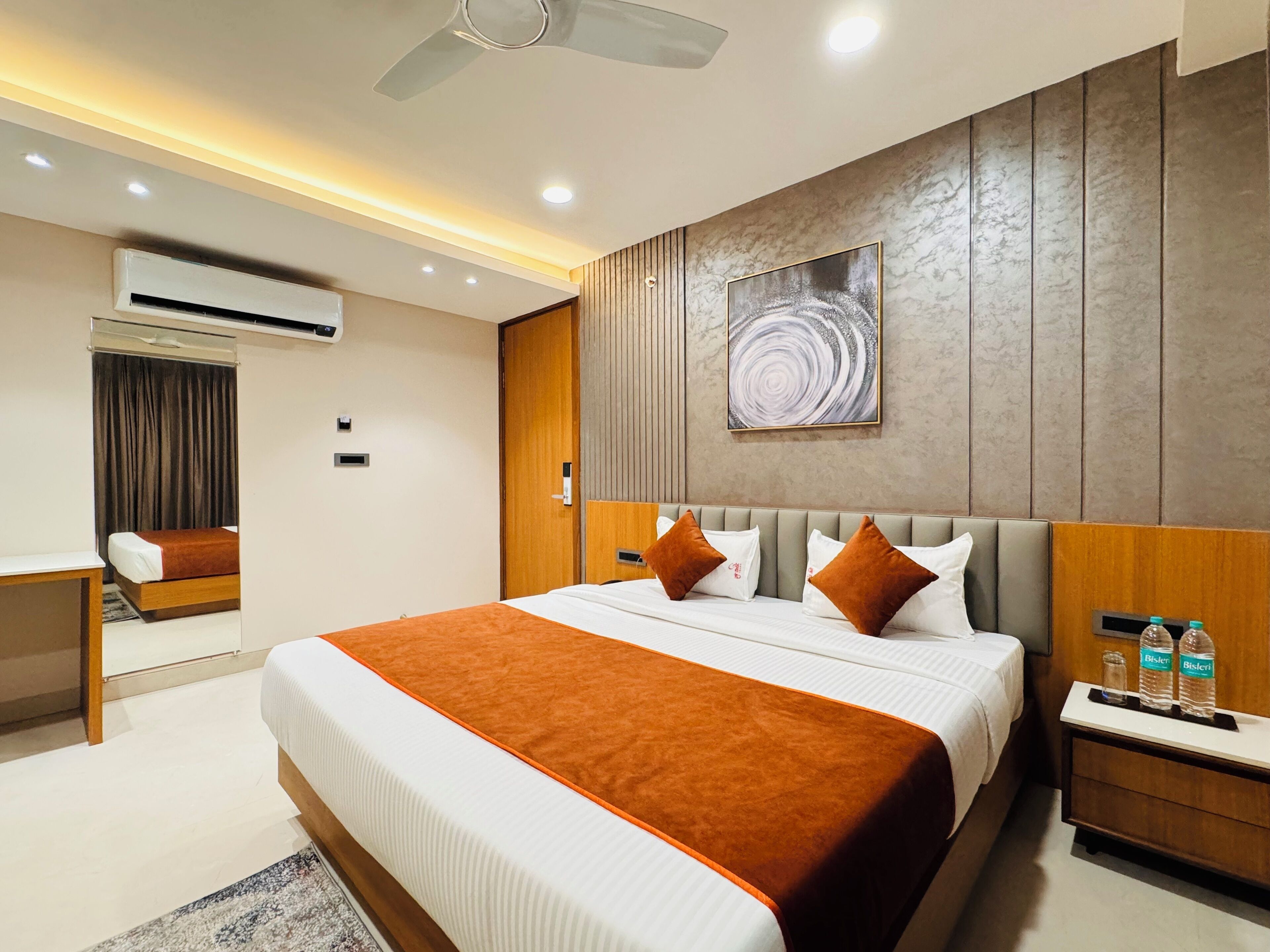 A modern hotel room with a large bed, a wooden headboard, a ceiling fan, an air conditioning unit, a bedside table with water bottles, and a large abstract painting on the wall.