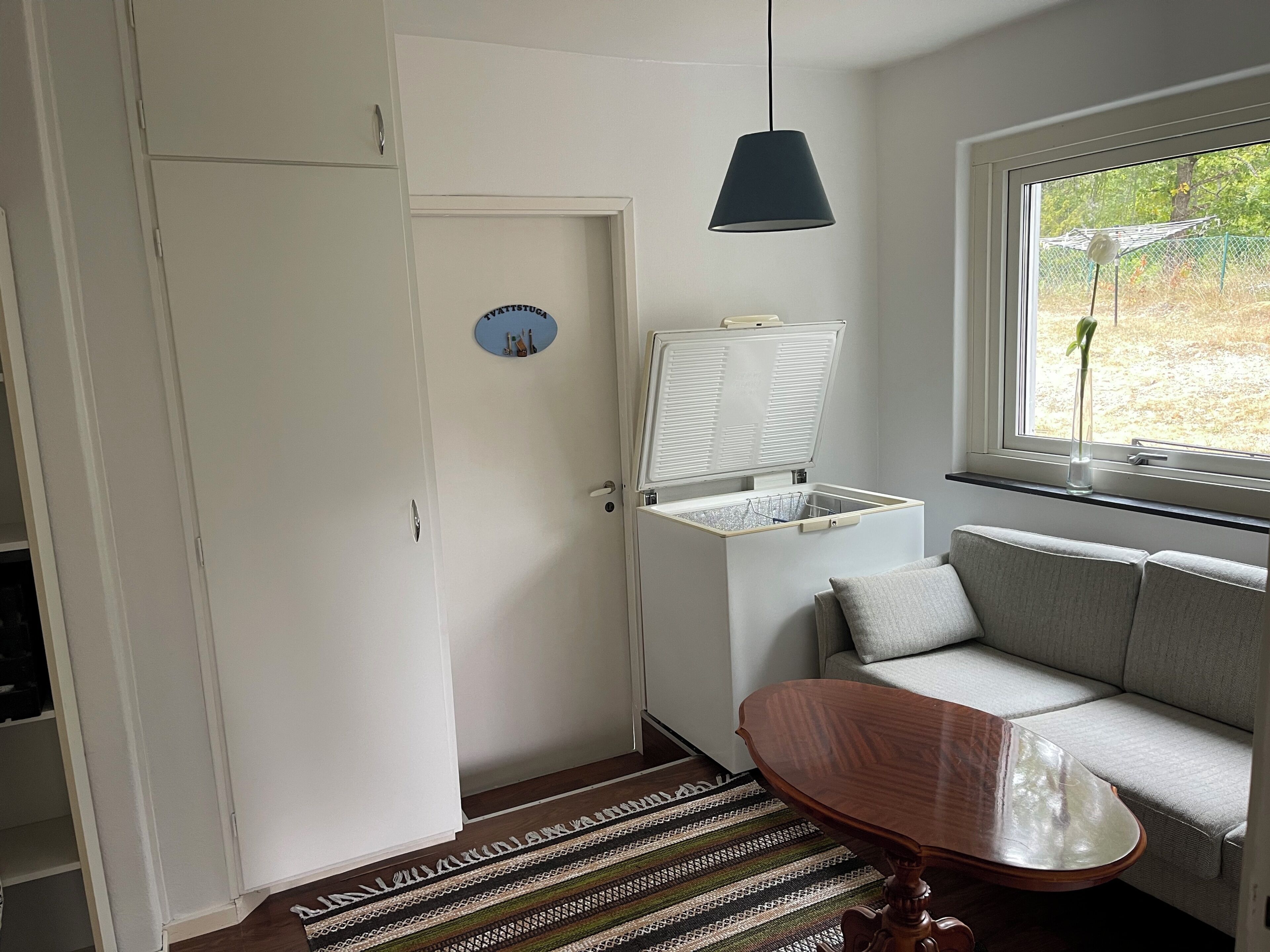A room with a white door, a small table, a sofa, a window with a view of greenery, and a ceiling light.
