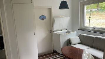 A room with a white door, a small table, a sofa, a window with a view of greenery, and a ceiling light.
