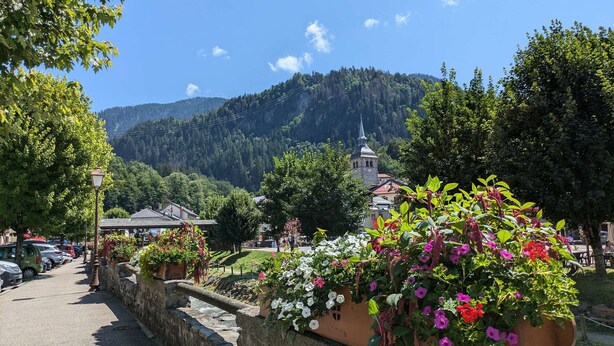 Un village pittoresque, avec des murs de pierre, des jardinières fleuries et une église surmontée d’une flèche.