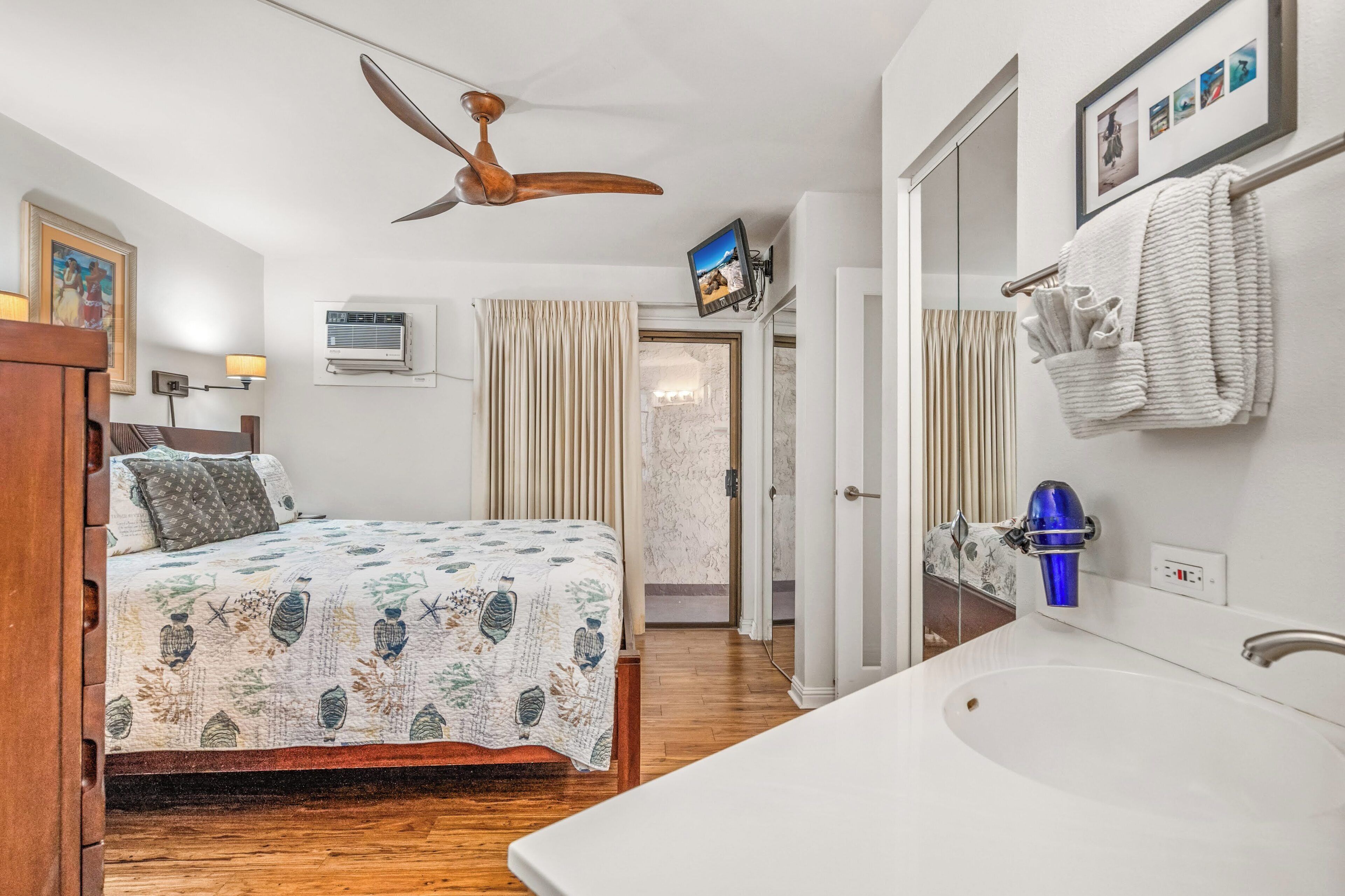A hotel room with a bed, a large window, a ceiling fan, a bathroom with a sink and mirror, and a wall-mounted TV.
