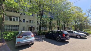 A parking area with several cars, a building with multiple windows and balconies, and trees providing shade.