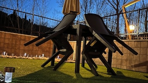 Une sculpture représentant des chaises aux formes abstraites, placée dans un jardin, avec un ciel dégagé et un mur de briques en arrière-plan.