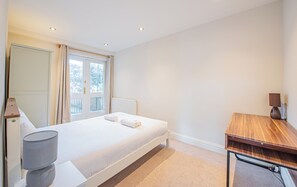 Ein Schlafzimmer mit Bett, Schreibtisch, Lampe und einem Fenster mit Vorhängen.