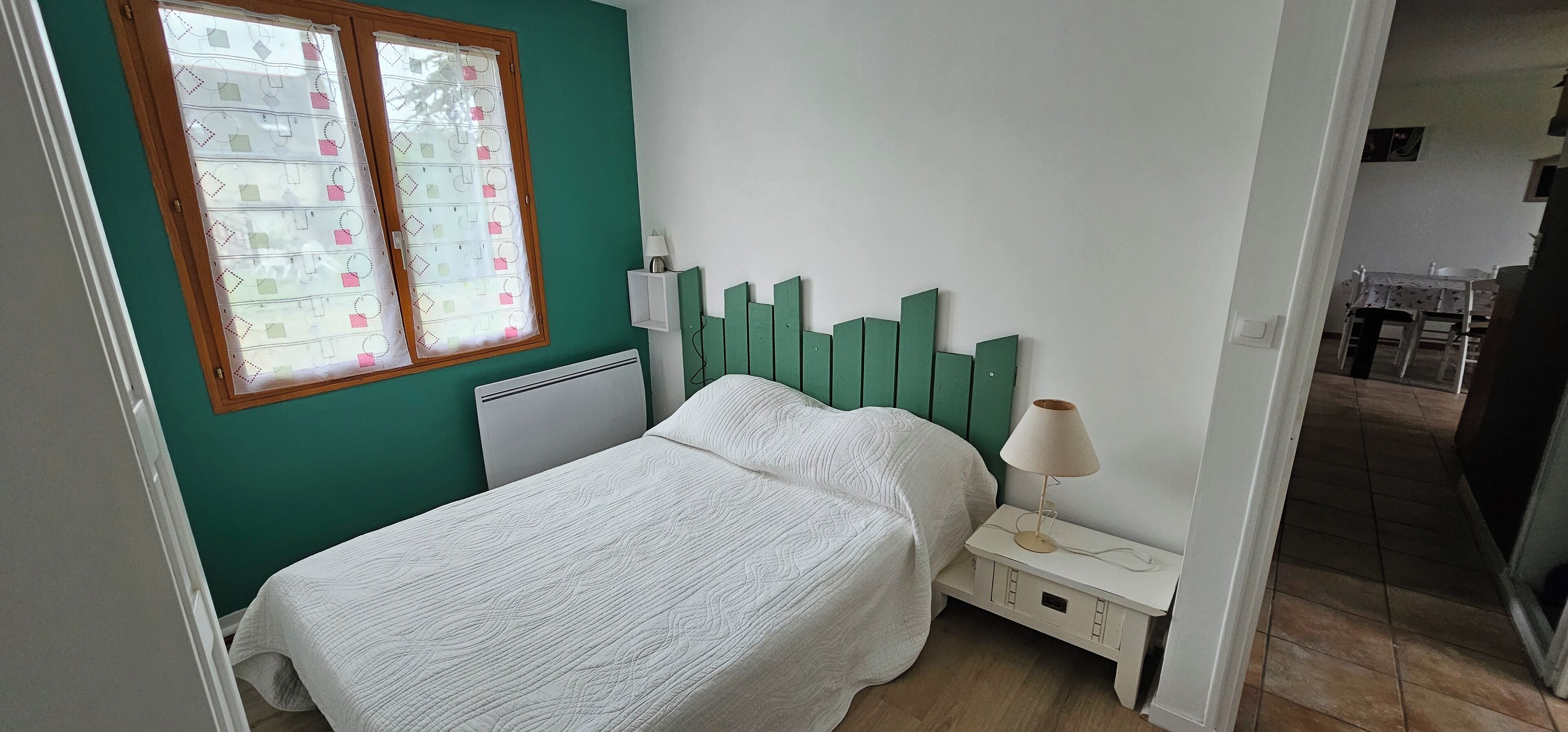 A bedroom with a bed, a nightstand with a lamp, a window with a curtain, and a radiator.