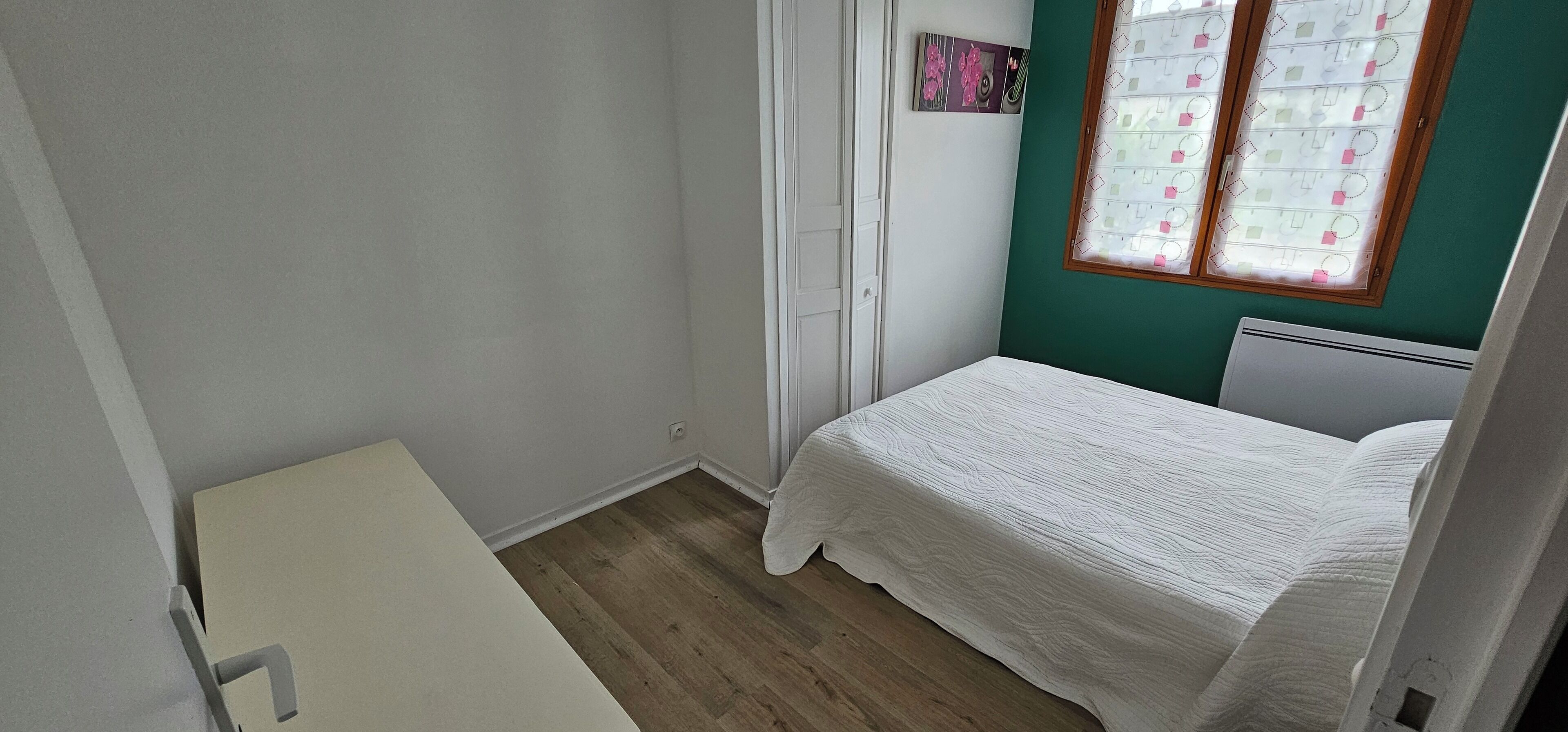 Une petite chambre à coucher, bien rangée, avec un lit simple, un bureau blanc et une fenêtre agrémentée d’un rideau à motifs.
