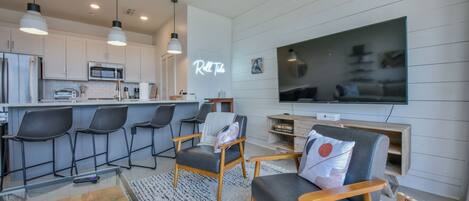 Una cocina moderna con gabinetes blancos, un microondas y un tostador. Hay una zona de bar con taburetes y un letrero de neón que dice "Roll Tide".