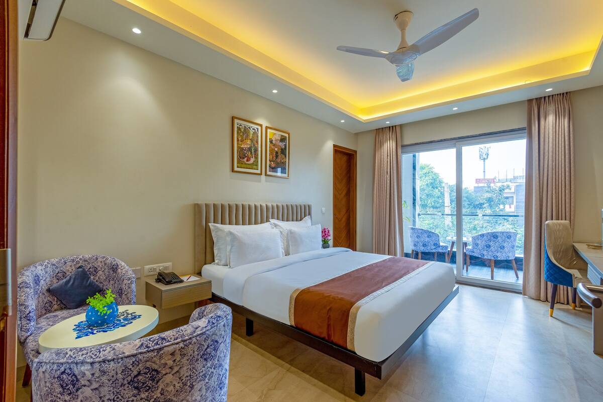 A hotel room with a bed, a desk, a chair, a ceiling fan, and a view of the outside.
