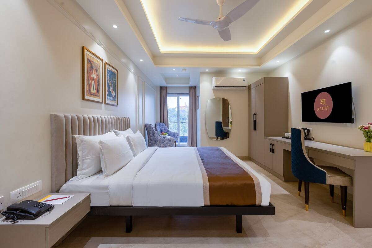 A modern hotel room with a large bed, a desk, a chair, a TV, and an air conditioning unit.