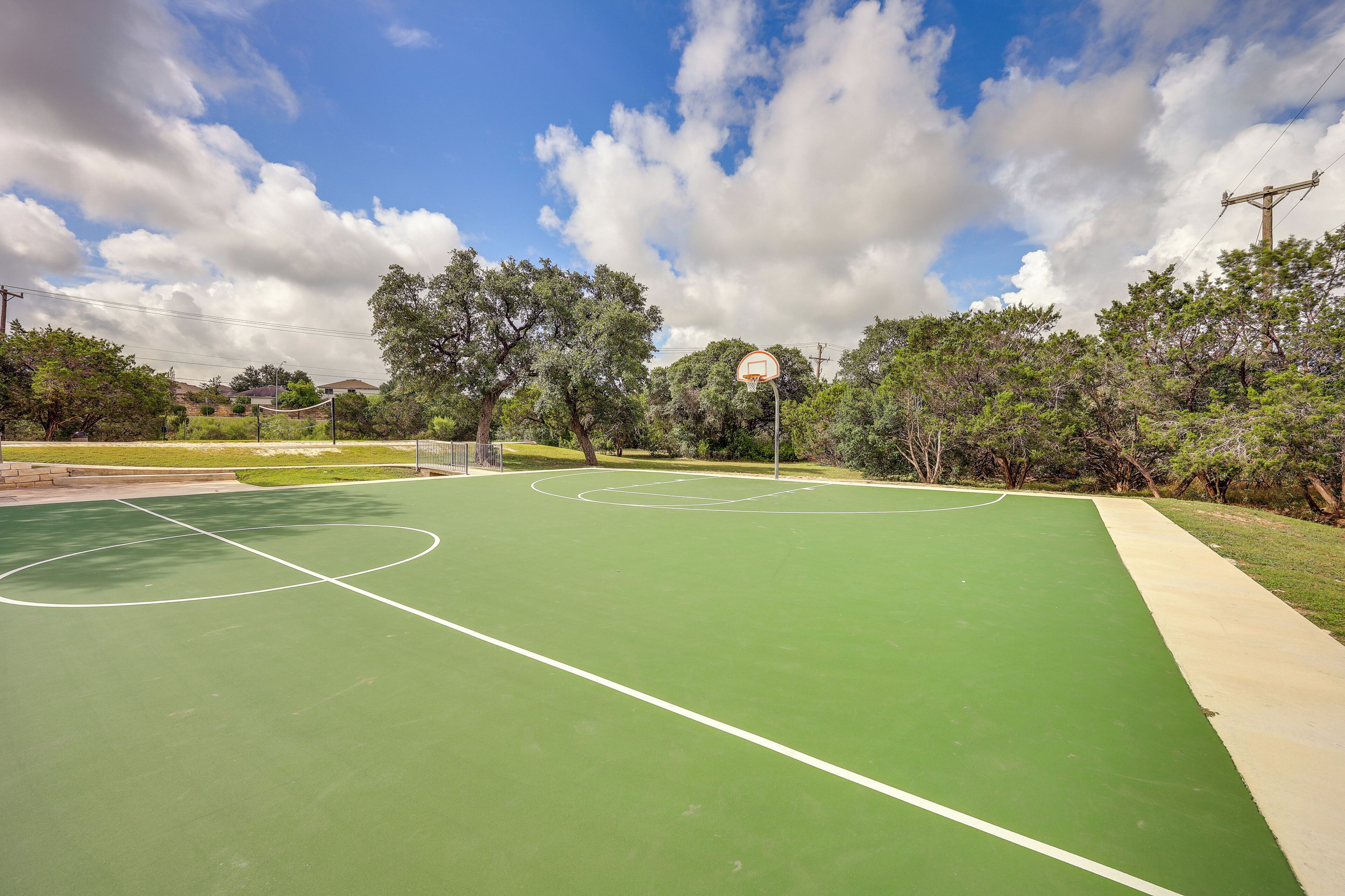 An outdoor basketball court with a green surface, surrounded by trees and a clear sky.