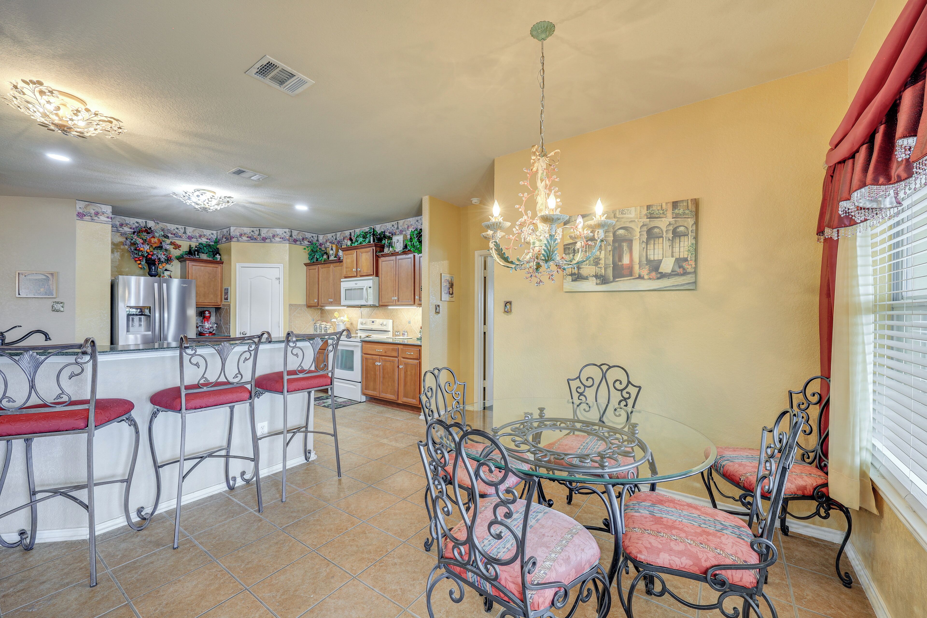 A dining area with a glass table and metal chairs, a kitchen in the background, and a chandelier.