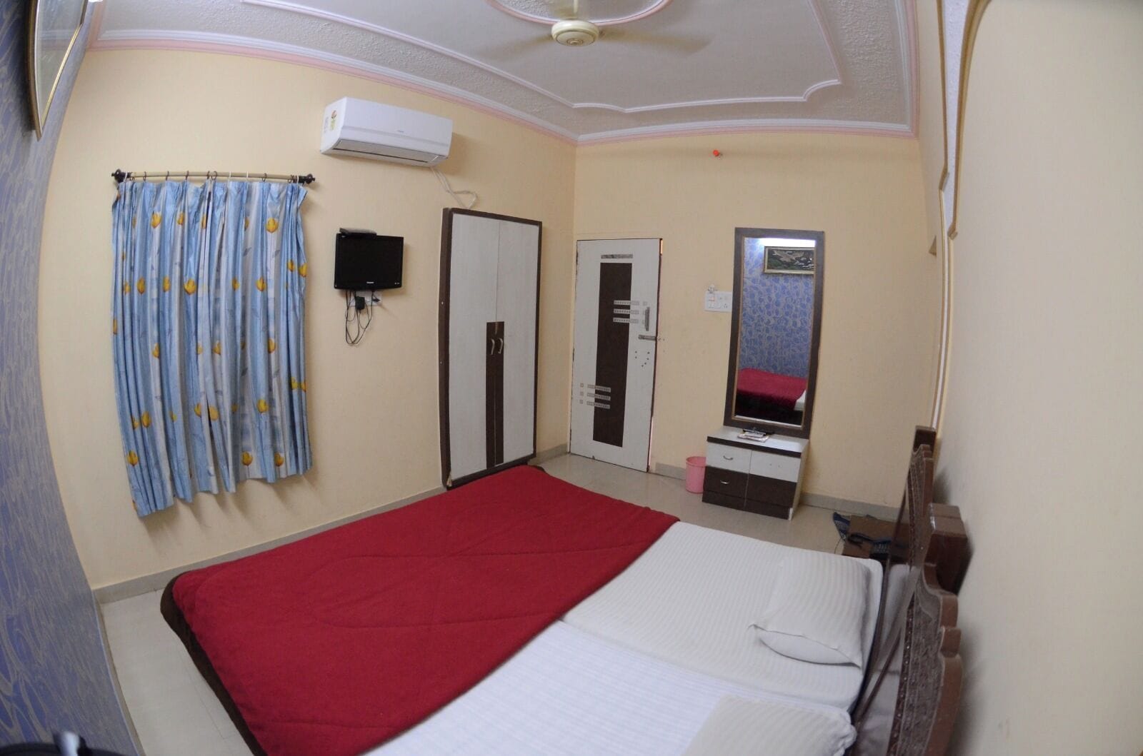 A bedroom with a bed, a red blanket, a ceiling fan, an air conditioner, a wardrobe, a mirror, and a curtain with a pattern.