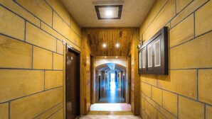Een smalle gang met houten wanden en een betegelde vloer, die leidt naar een lichte kamer met uitzicht op een gebouw.
