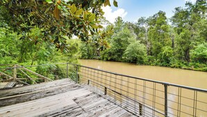 Uma ponte de madeira com grades de metal que leva a um rio, cercado por vegetação densa e verde.