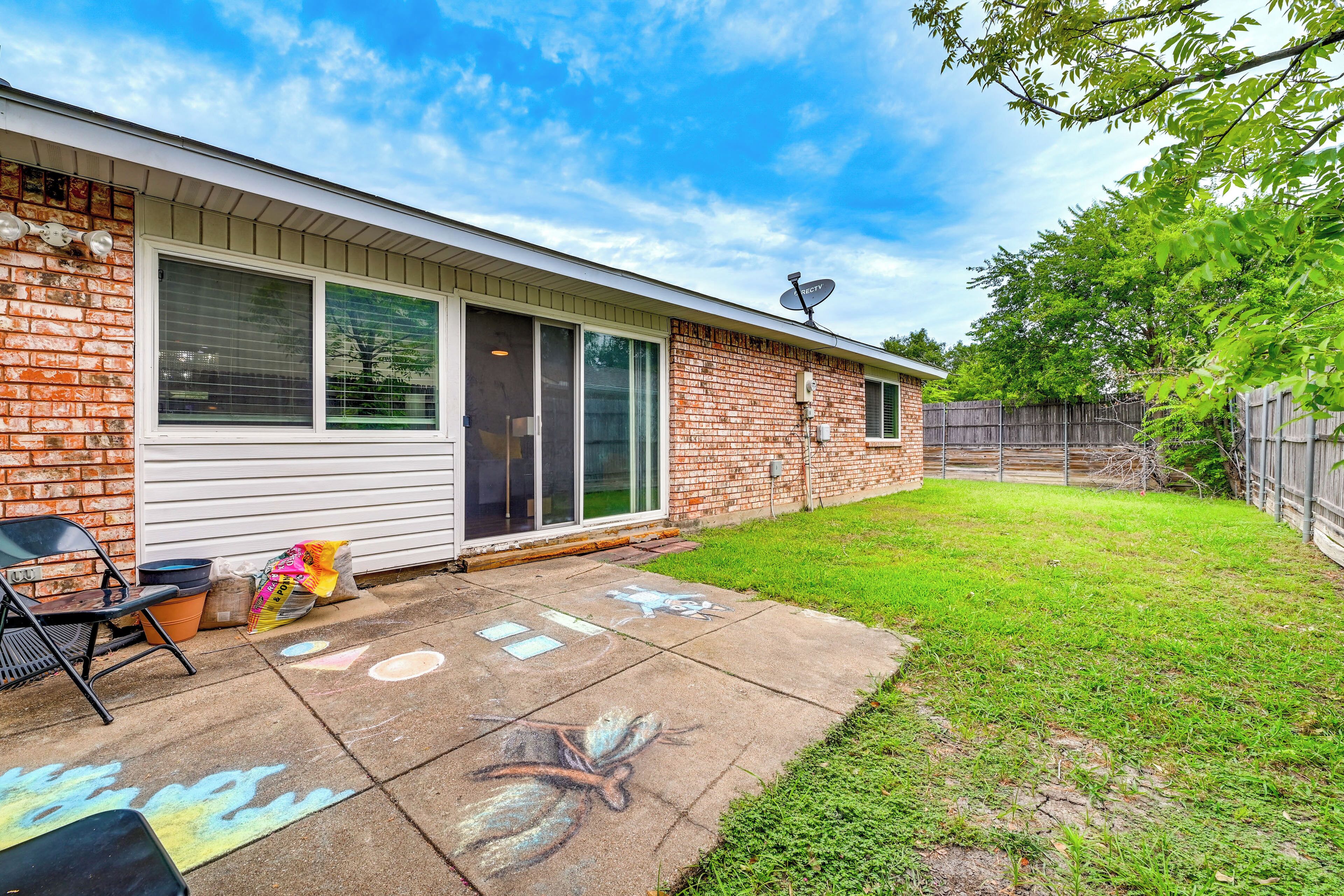 A residential backyard with a brick house, a sliding glass door, a patio with a chair, and a grassy lawn.