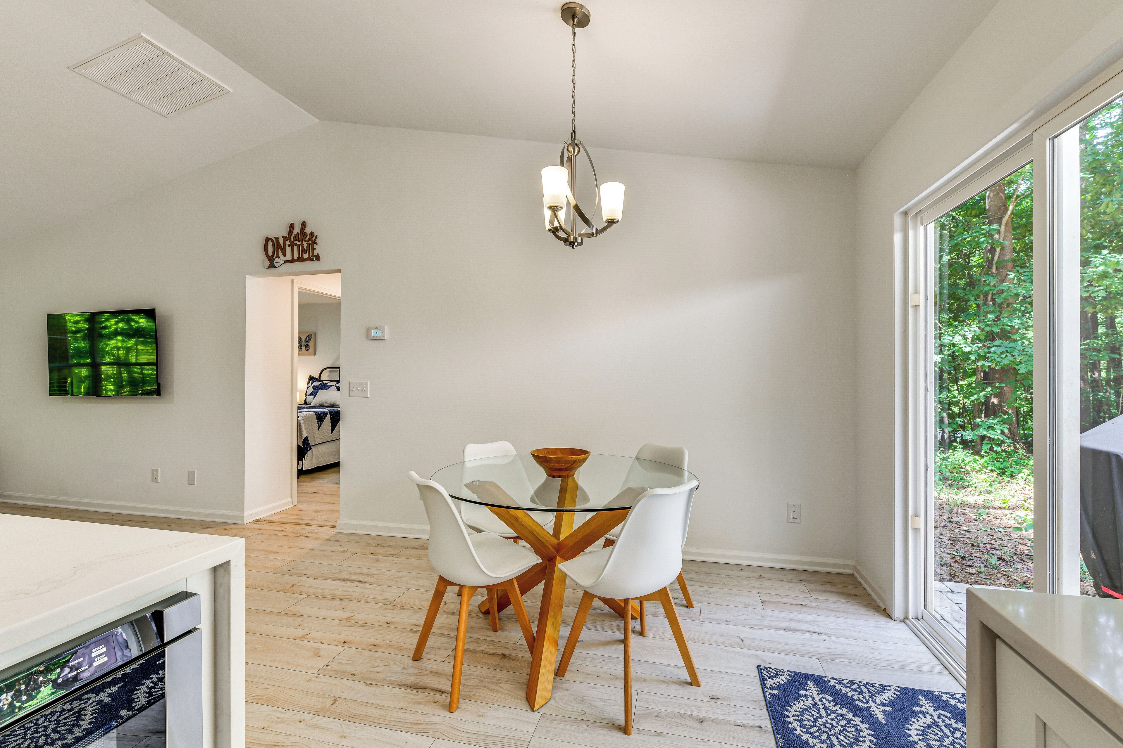 A modern dining area with a glass table and white chairs, a chandelier, and a view of greenery outside.
