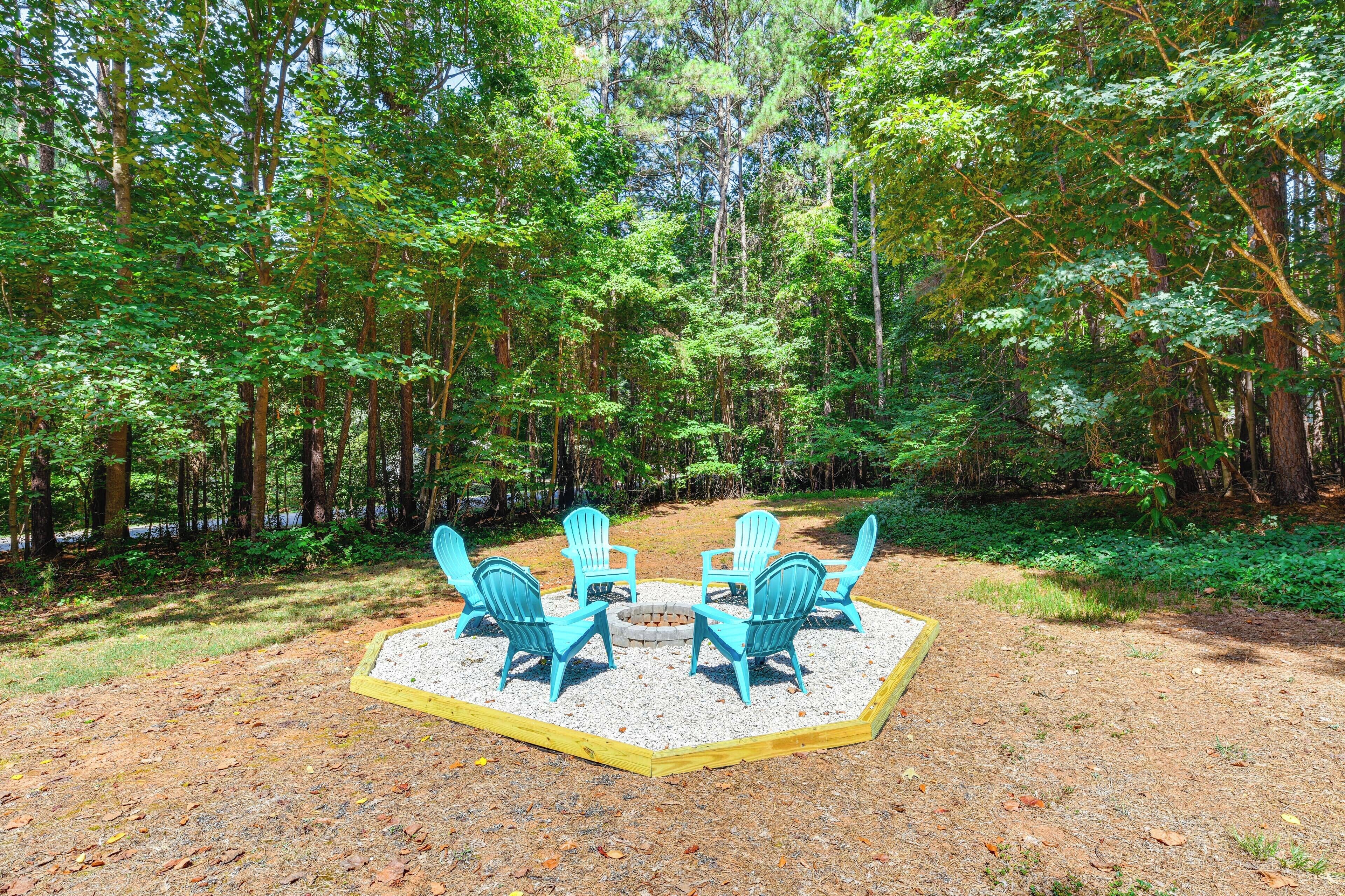 A forested area with a fire pit and four blue chairs arranged in a circle.