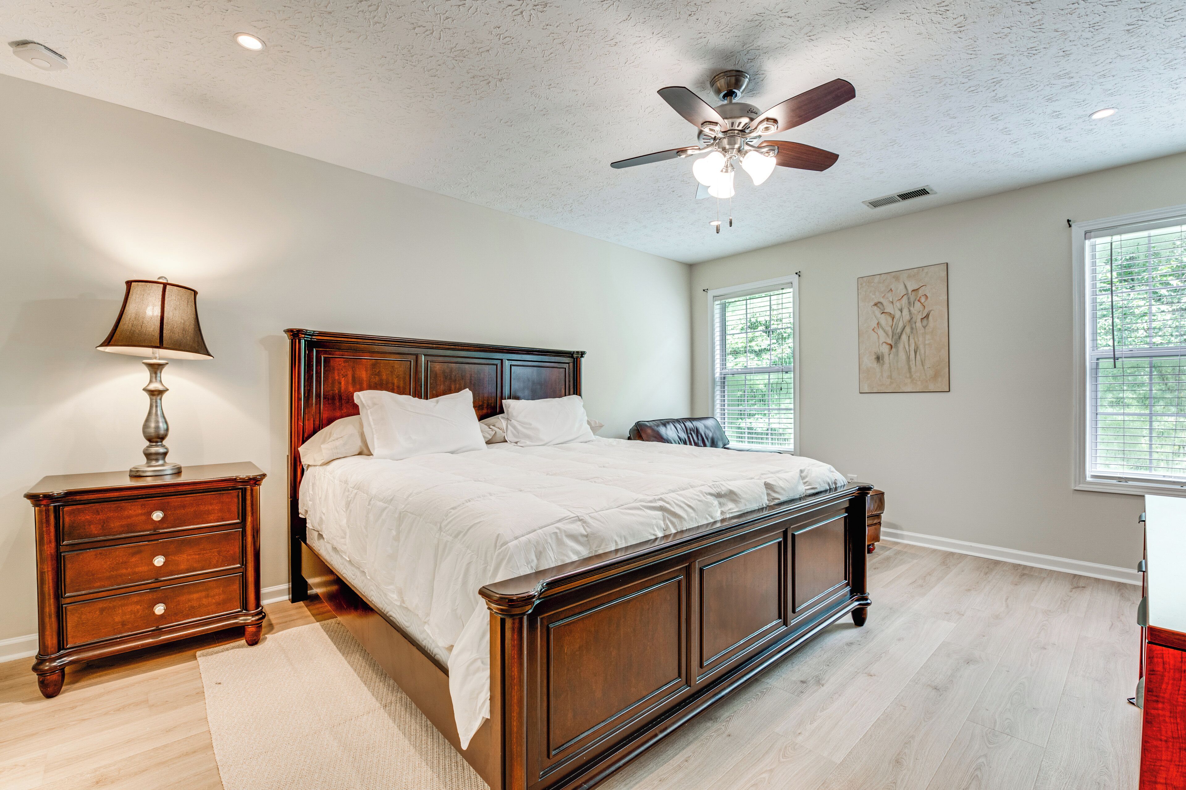 A bedroom with a wooden bed, a nightstand with a lamp, a ceiling fan, and a window with blinds.