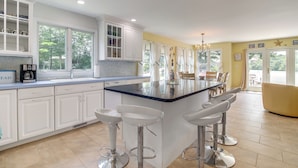 A modern kitchen with white cabinets, a large island with bar stools, and a view of the outdoors through large windows. - Plymouth Getaway w/ Private Lake Beach + Kayaks! (Plymouth)