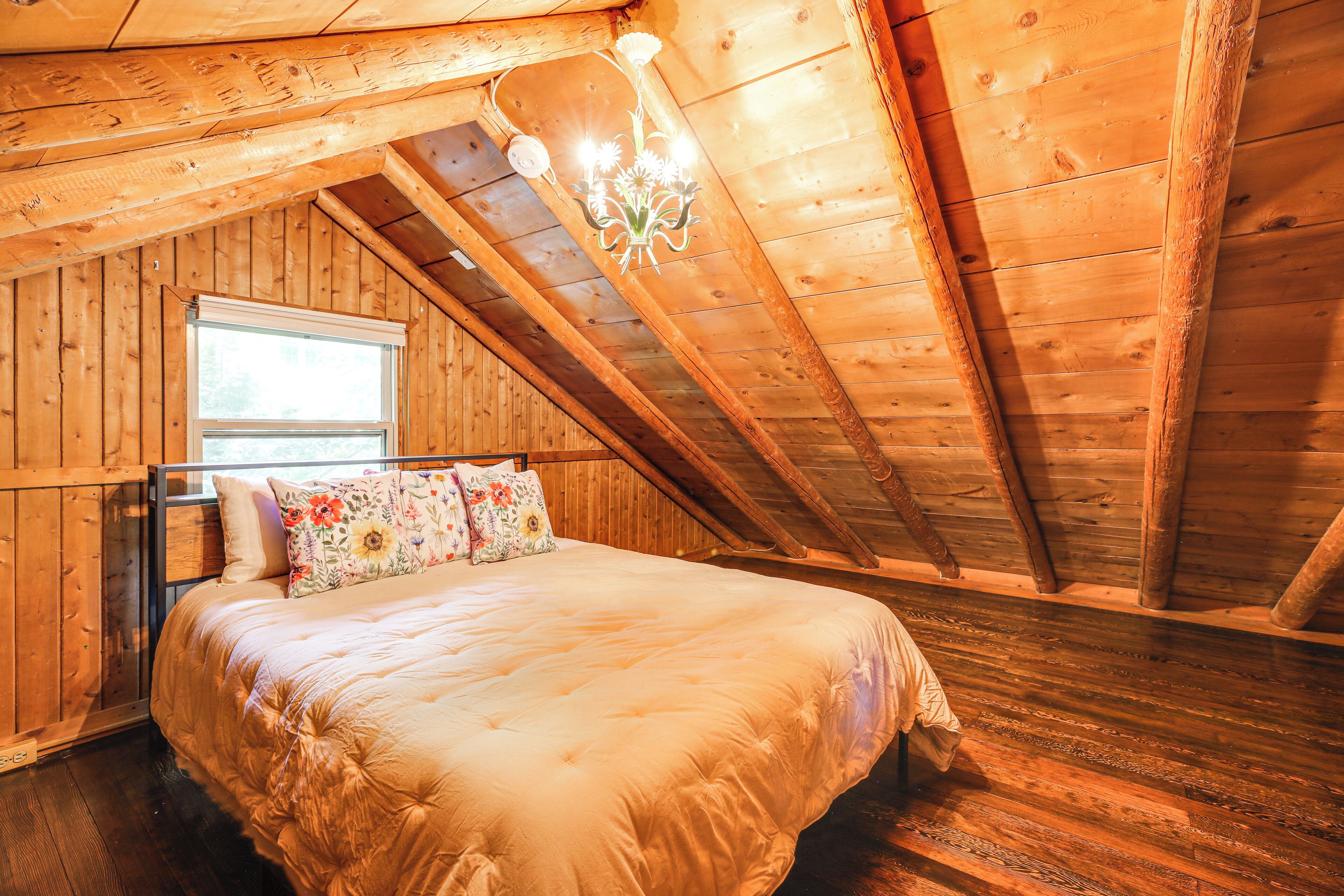 A cozy attic bedroom with a wooden ceiling, a large bed with floral pillows, and a window providing natural light.
