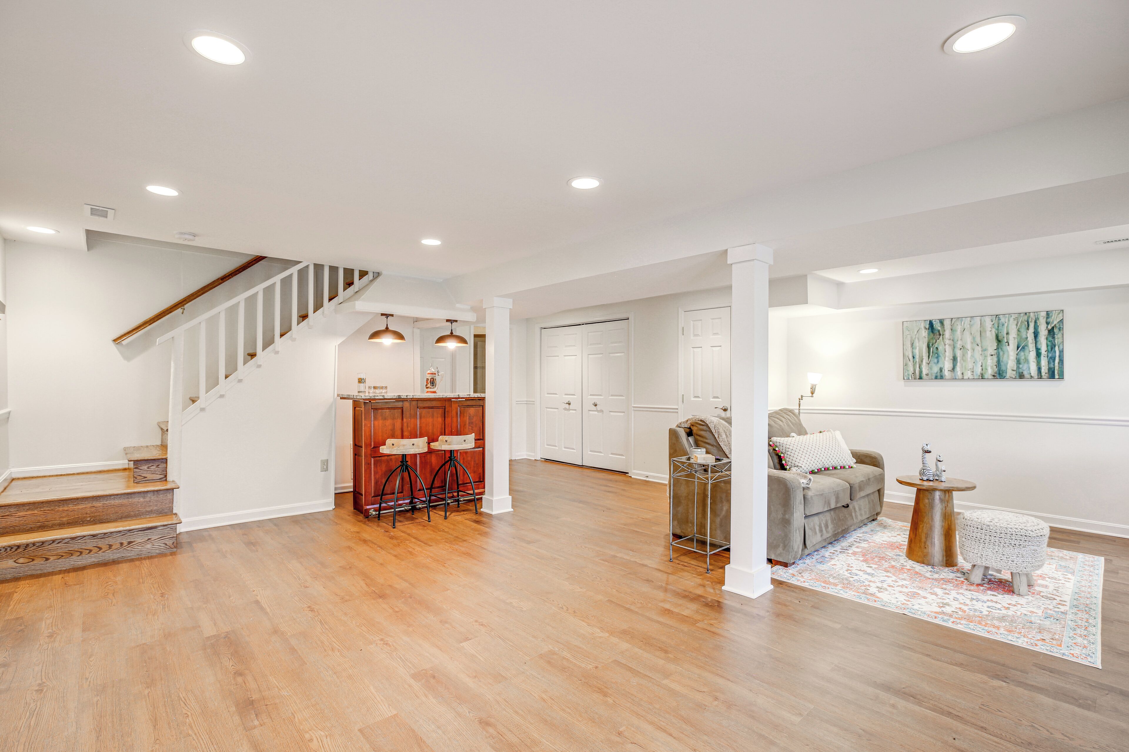 A spacious living area with hardwood flooring, a staircase, a kitchen area with bar stools, a gray sofa, and a round wooden table.