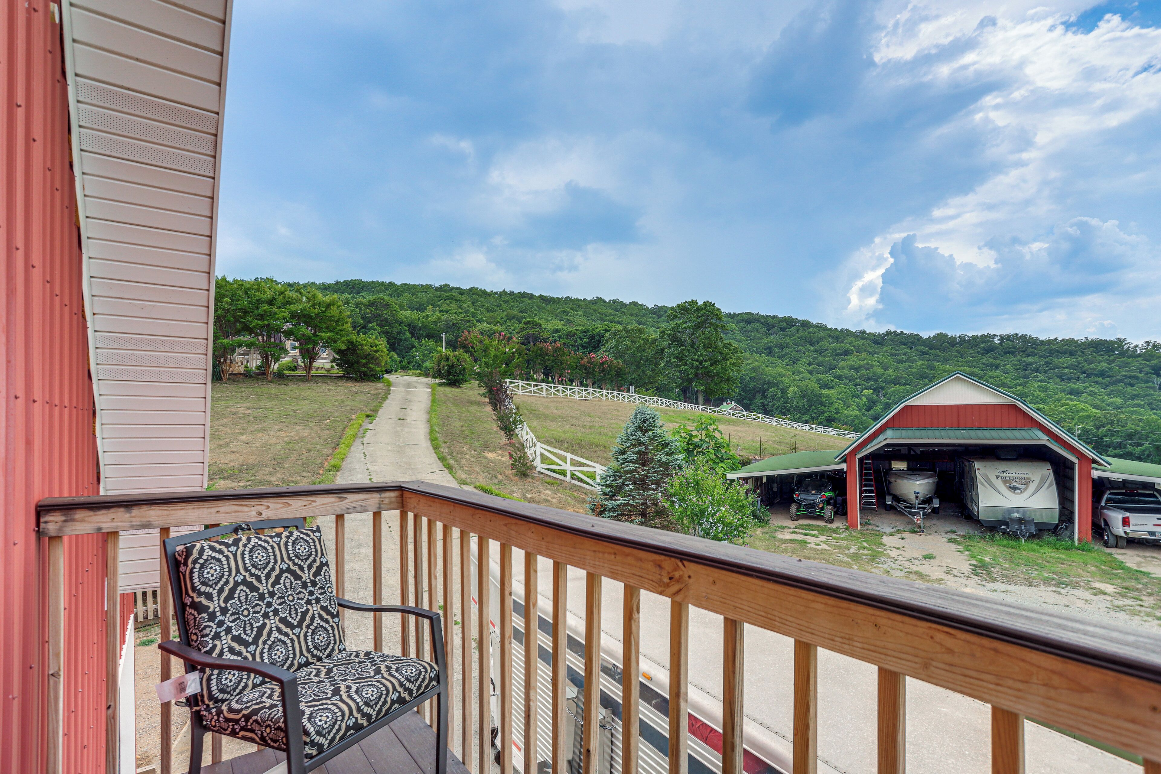 A porch with a chair, a red barn, and a trailer in the background.
