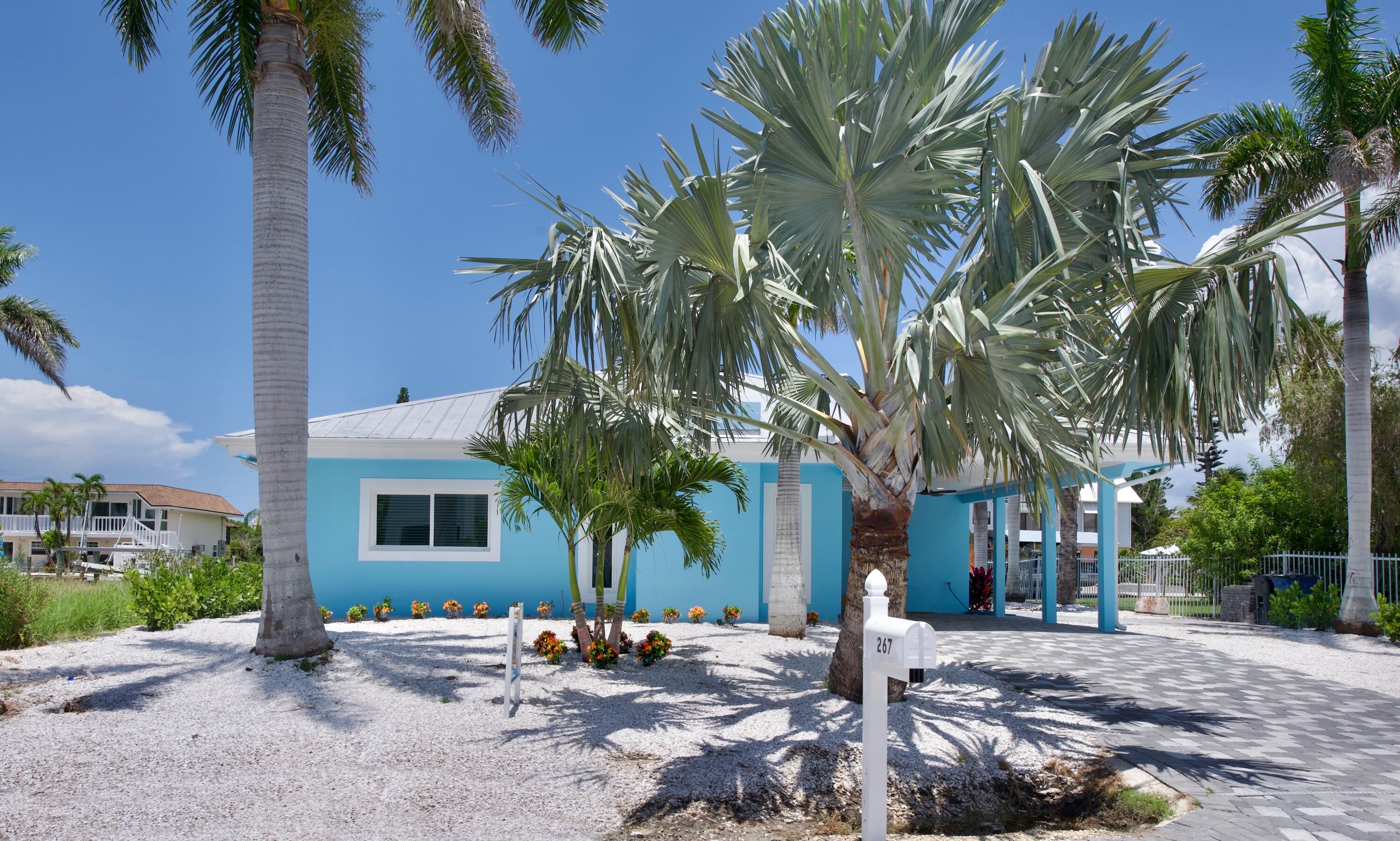 A house with a blue exterior, surrounded by palm trees and a gravel driveway.
