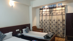 A hotel room with two beds, a wooden headboard, a bedside table with a glass, and a floral curtain. - HOTEL SHILLONG RAINS INN BY ROH (Shillong)