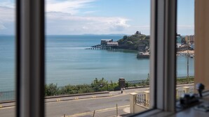 A coastal view from a window, featuring a pier extending into the sea, a hilltop structure, and a road with a fence and greenery in the foreground. - Nash House - Seafront With Parking Close to Town (Tenby)