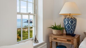 A room with a large window offering a view of the sea and a pier. There is a wooden side table with a lamp and a potted plant. - Nash House - Seafront With Parking Close to Town (Tenby)