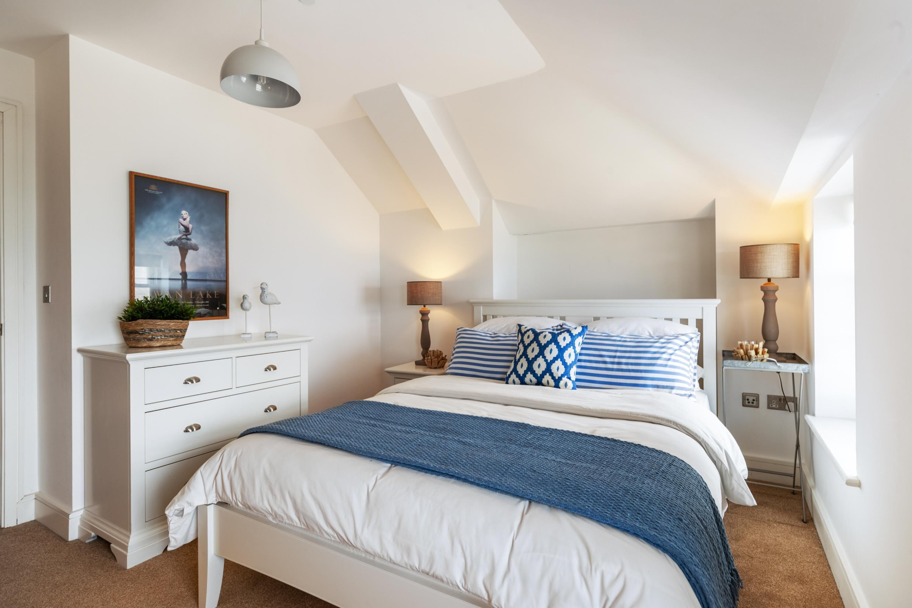 A bedroom with a white bed, blue and white bedding, a nightstand with a lamp, and a wall-mounted shelf with a plant.