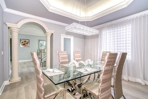 Een eetruimte met een glazen tafel, beige stoelen, een kroonluchter en uitzicht op een trap.