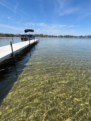 Un lago cristalino con un muelle que se adentra en el agua, un bote amarrado al muelle, y una orilla distante con edificios.