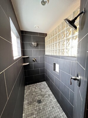 Un baño pequeño con azulejos oscuros, una ventana, un cabezal de ducha y un estante en la pared.