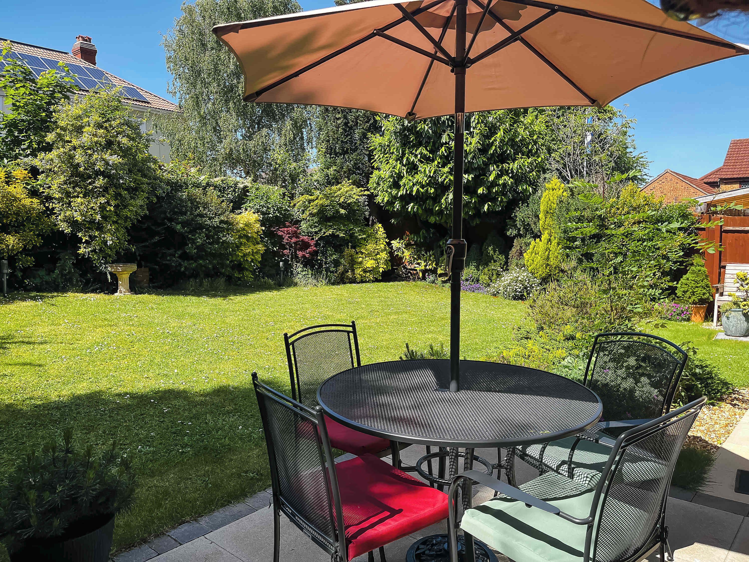 Une terrasse avec une table ronde et des chaises, un grand parasol, et un jardin avec différentes plantes et un abreuvoir pour oiseaux.
