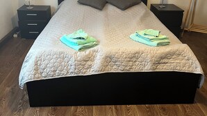 Una cama bien hecha con cubrecama acolchado, dos almohadas y toallas dobladas sobre un suelo de madera, con una mesita de noche y una lámpara a cada lado.