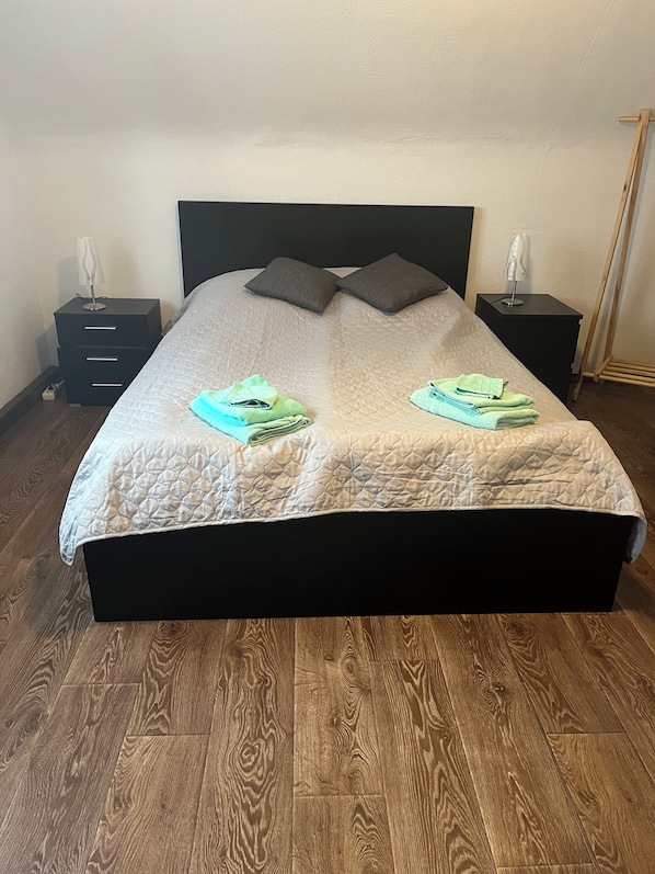 Una cama bien hecha con cubrecama acolchado, dos almohadas y toallas dobladas sobre un suelo de madera, con una mesita de noche y una lámpara a cada lado.