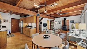 A spacious living area with a wooden ceiling, a dining table, and a sofa. A kitchen with modern appliances is visible in the background.