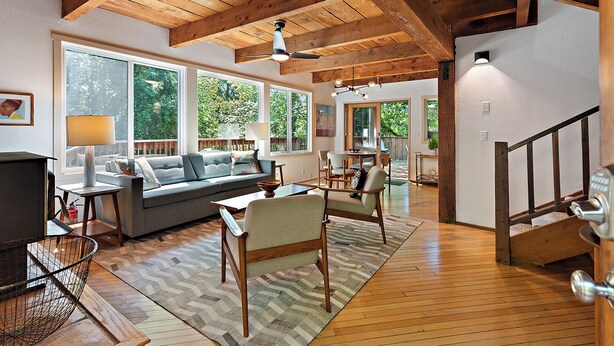 A spacious living room with a wooden ceiling, large windows, a sofa, a coffee table, and a dining area.