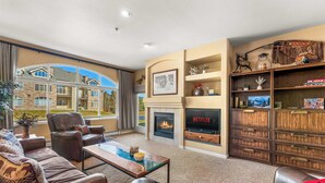 A living room with a fireplace, a bookshelf, a leather sofa, and a large window. - Beaver Lodge — 2BR Frisco Condo, Walk to Lake Dillon, Hot Tub, Sleeps 5 (Frisco)