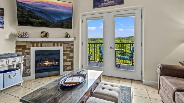A living room with a fireplace, a large painting, and a balcony with chairs. - Top Rated Condo | Mtn Views- POOL- Playground (Pigeon Forge)