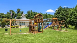 A playground with wooden structures, including a slide and swings, set against a backdrop of trees and a building. - Top Rated Condo | Mtn Views- POOL- Playground (Pigeon Forge)