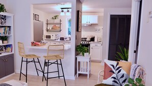 Cocina moderna con armarios blancos, microondas y estufa. Hay dos sillas de mimbre y una mesita pequeña con una planta.